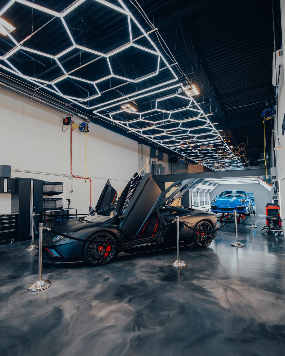 Lamborghini roadster with doors open beside a Porsche GT3 RS inside ZAutoCare under hexagonal LED lighting